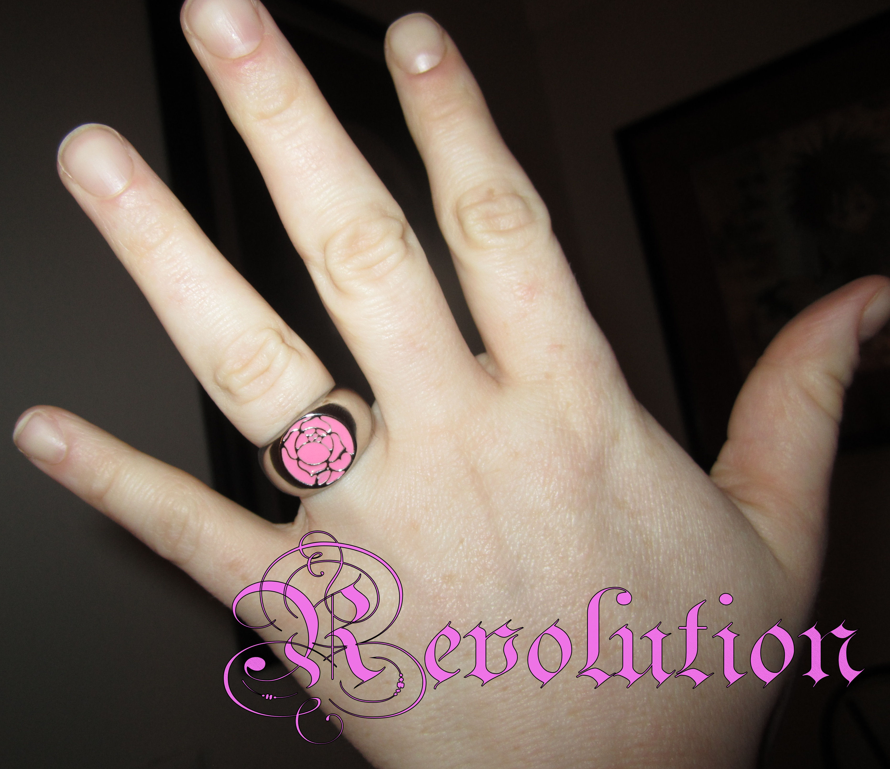 A photo of my hand. I am wearing the Rose Crest ring on my ring finger, and the image says "Revolution" in stylized font.